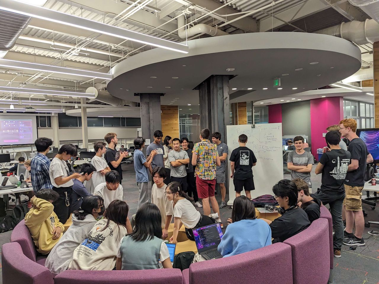 A large group of people gathered around a whiteboard in a makerspace. At the bottom of the image is a purple couch arranged in a half-circle, which many are sitting on.