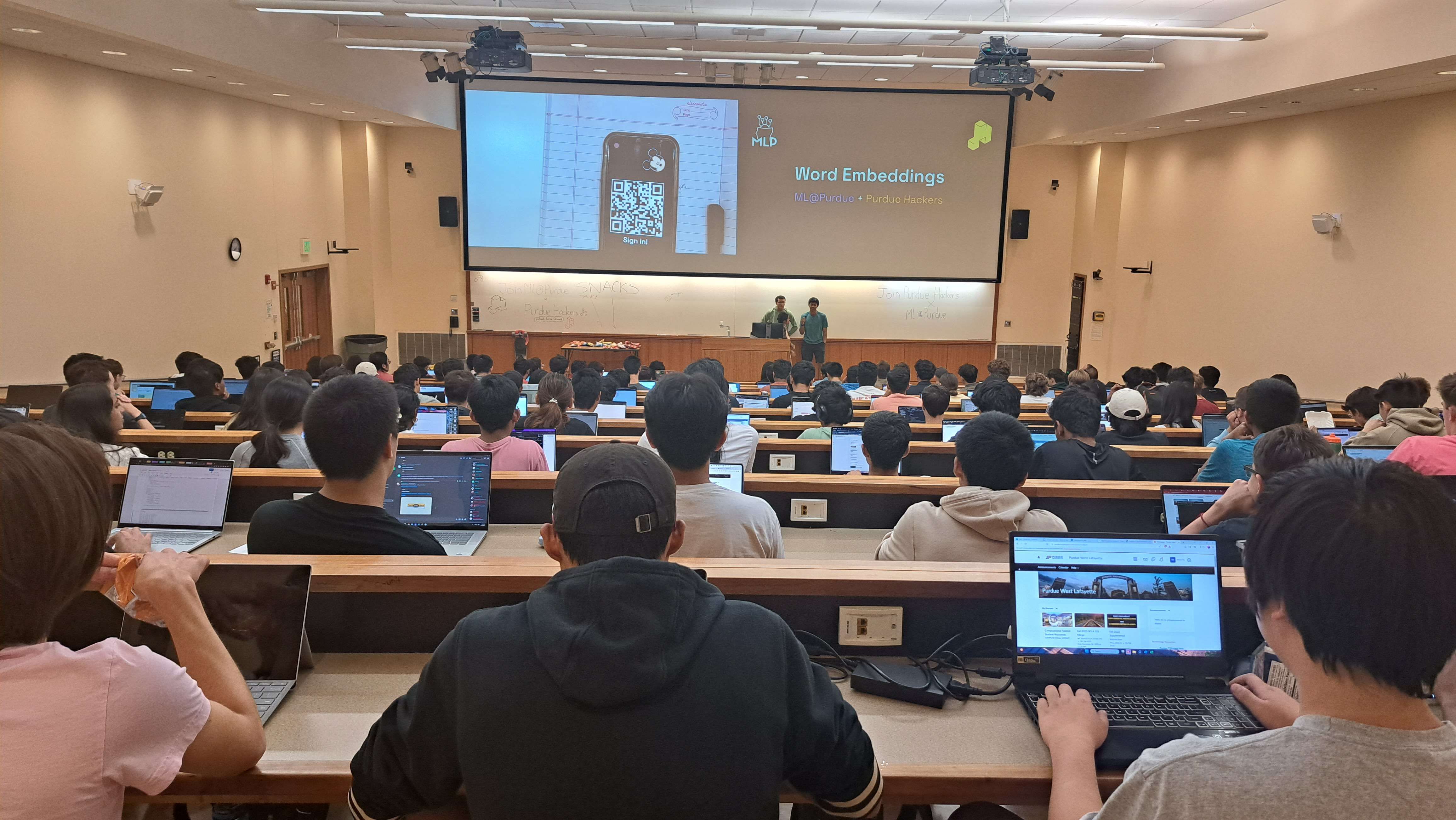 A picture from the back of a lecture hall. Every seat is full of students, and at the front of the room a small group of students are presenting a workshop on word embeddings